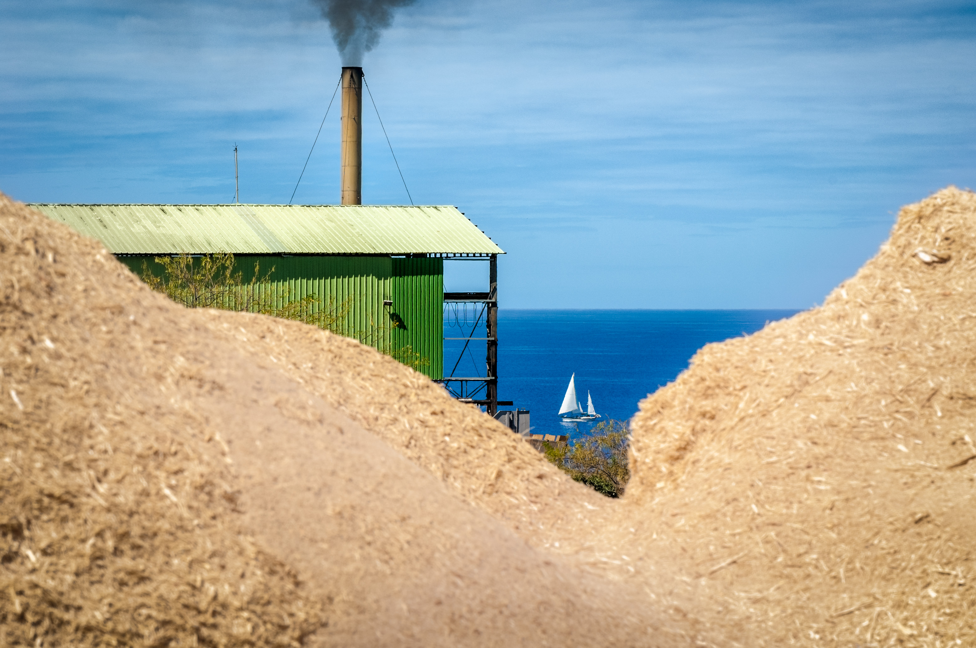 France, Caraïbes, Petites Antilles, Guadeloupe, Basse-Terre, Distillerie de rhums agricoles Bologne, Un voilier passe au loin, entre 2 tas de bagasse, le résidu fibreux de la canne à sucre qu'on a passée par le moulin pour en extraire le suc // France, Caribbean, Lesser Antilles, Guadeloupe, Basse-Terre, Distillery of agricultural rum Bologna, A sailboat passes away, between 2 heaps of bagasse, the fibrous residue of sugar cane that has passed through the mill to extract the juice